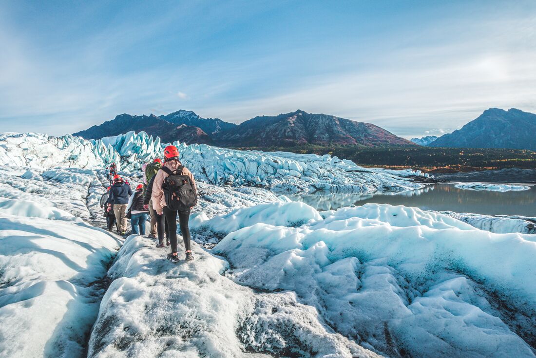 Column of travellers in safety gear hike on the icey Matanuska Glacier with Alaskan landscape in the distance