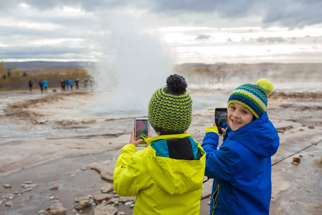 Young travellers smile and watch as Strokkur geyser erupts in front of them as they film on their phones
