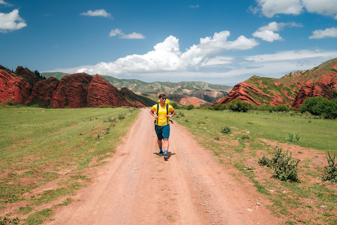Traevller in yellow hikes in the red and green landscape of Jeti Oguz with clear sunny clouds in background