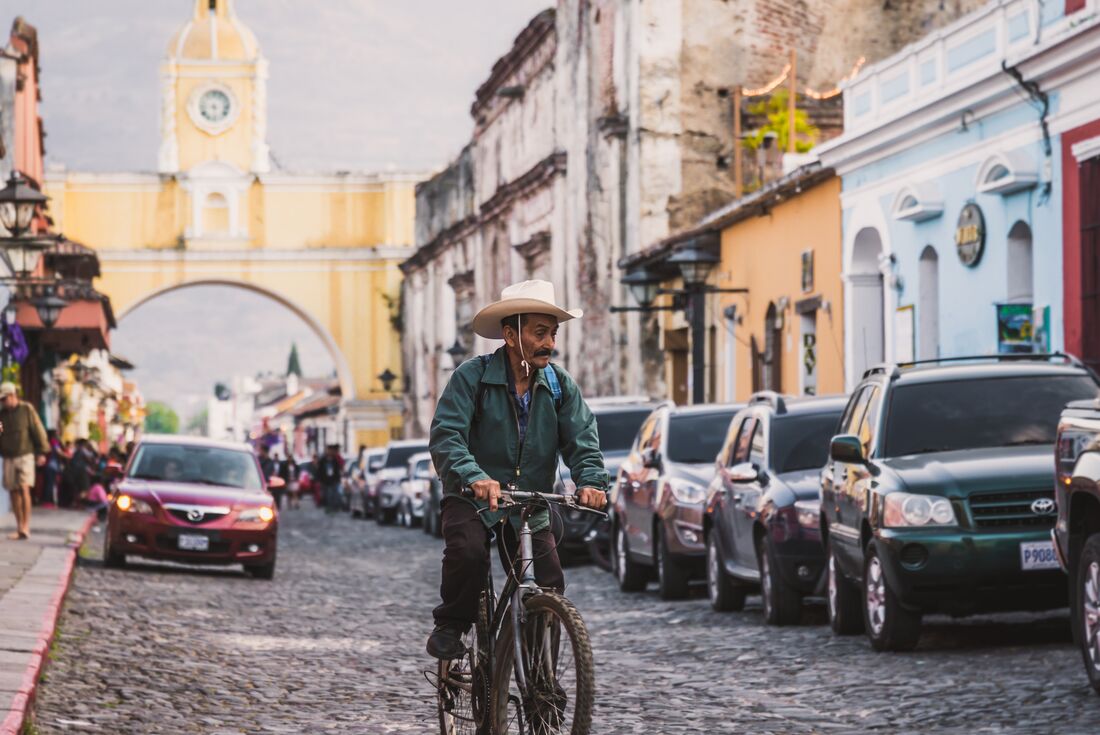 Local man in wide brimmed hat riding a mouuntain bike down the streets of Antigua with old buildings and new cars around